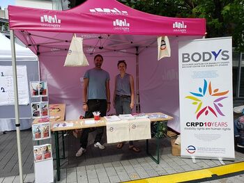Infostand der EvH RWL. Hinter einem Tischen stehen Prof. Dr. Stefan Schache von BODYS und seine EvH-Kollegin Kristin Wallmeyer. Beide lachen fröhlich in die Kamera. Vorn rechts steht ein Aufsteller mit dem BODYS-Logo und der Schrift "10 years UNCRPD".