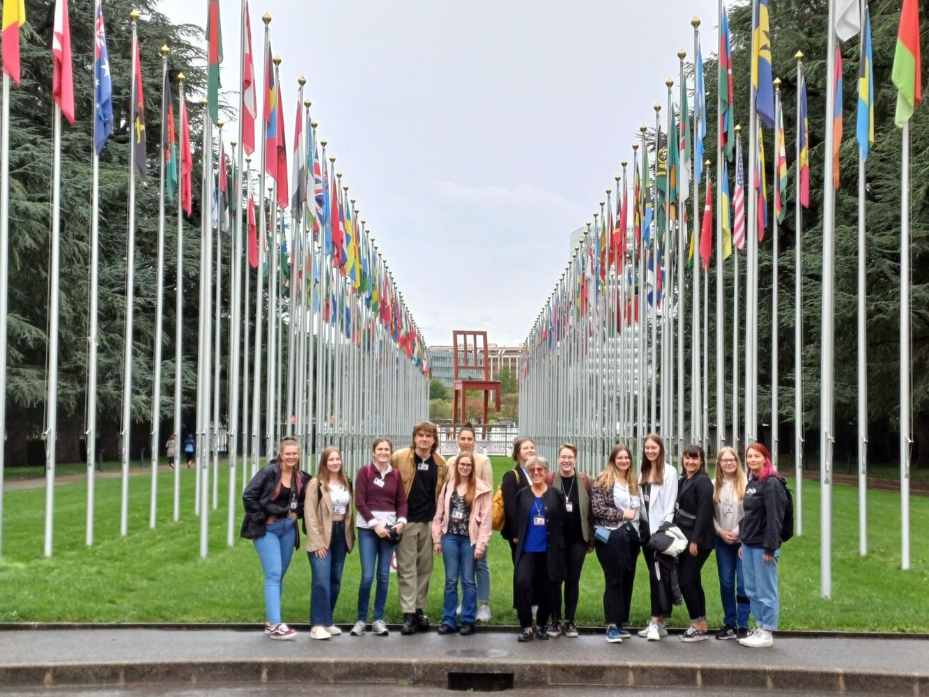 15 Studierende und Theresia Degener stehen vor dem Flaggenspalier der Vereinten Nationen in Genf, im Hintergrund die Plastik Broken Chair.