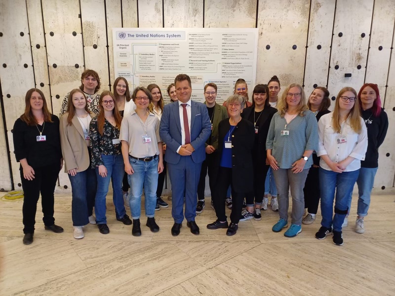 Eine große Gruppe von Studierenden, die Exkursionsleiterinnen und in der Mitte Jürgen Dusel. Im Hintergrund eine Infotafel zum System der Vereinten Nationen.