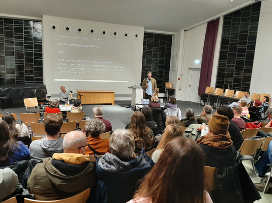 Viele Menschen in einem Hörsaal, auf der Bühne Udo Sierck sitzend und Stefan Schache stehend, im Hintergrund eine große Leinwand für Schriftdolmetschung