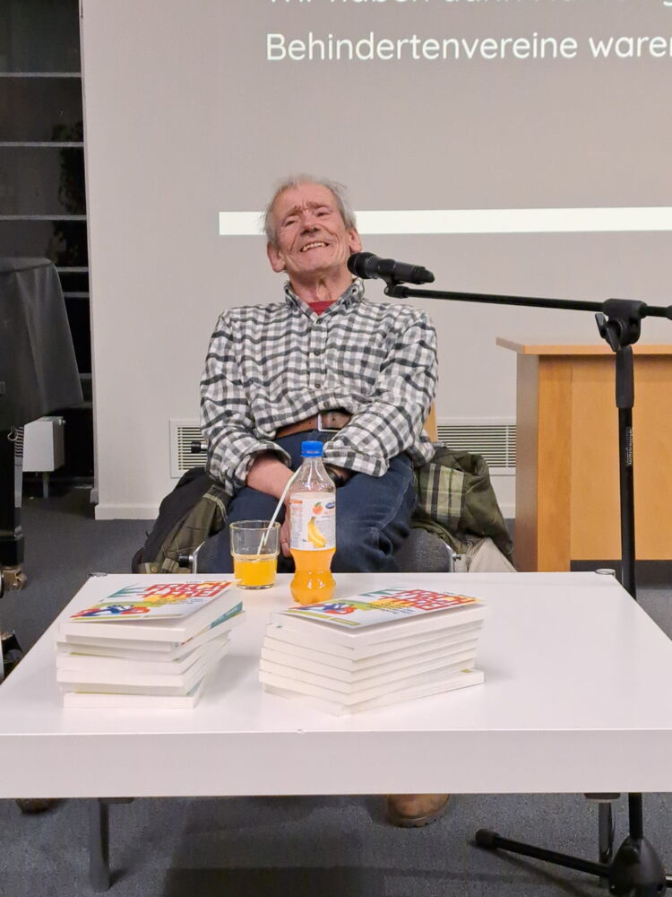 Udo Sierck lächelt in die Kamera. Er sitzt hinter einem flachen Tisch, auf dem zwei Stapel seiner Bücher liegen. Ein Mikrofon steht neben ihm.