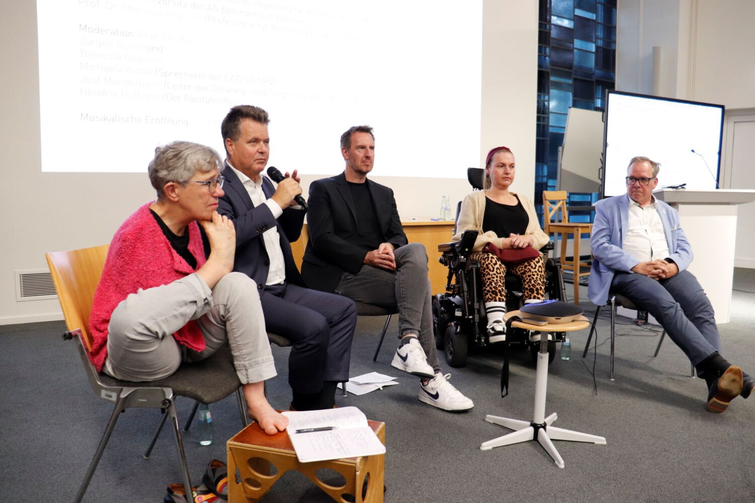 Auf einer Bühne sitzen eine armlose Frau, ein blinder Mann, eine Frau im Rollstuhl und zwei weitere Männer. Die Personen heißen: Theresia Degener, Jürgen Dusel, Hendrik Hoffjan, Michaele Kusal und Jost Manderbach.